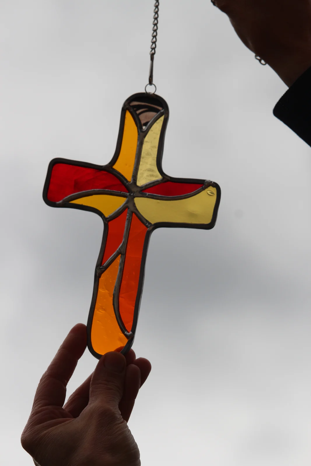 Croix à suspendre en vitrail tiffany aux nuances rouge, jaune et orange, tenue en extérieur devant l'herbe.