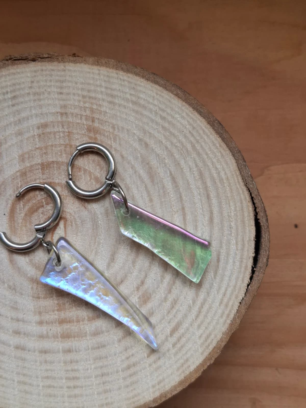 Boucles d'oreilles pendantes en verre aux reflets holographiques vert et violet sur un rondin de bois.