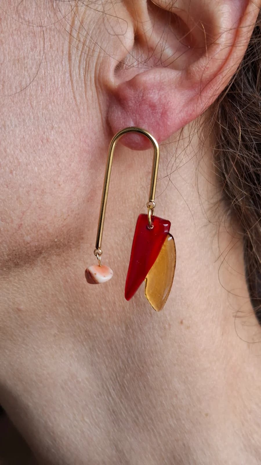 Boucle d'oreille graphique dorée portée à l'oreille, ornée de verre rouge, ambré et d'une perle.