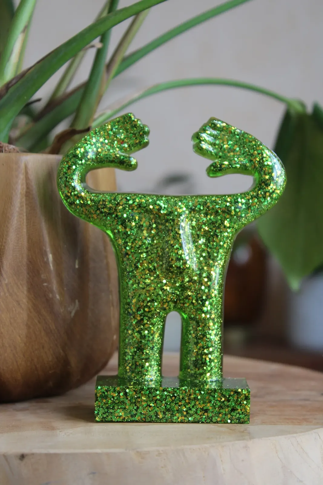 Statuette stylisée verte pailletée aux bras levés devant une plante d'intérieur.