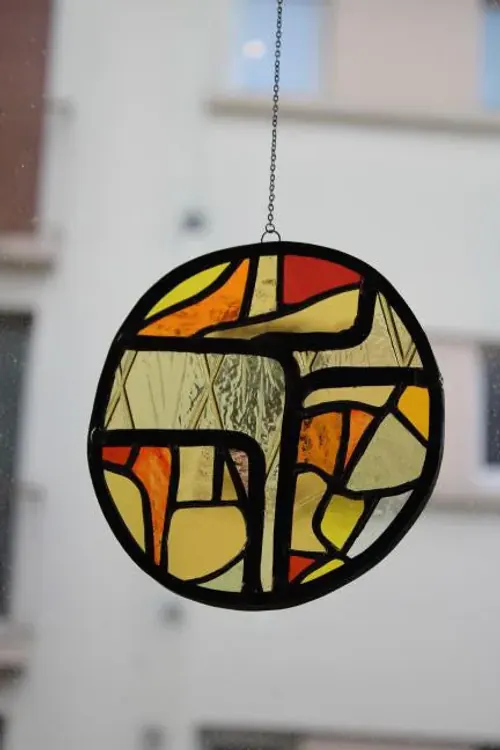 Vitrail rond abstrait aux tons chauds suspendu par une fine chaîne devant une fenêtre lumineuse.