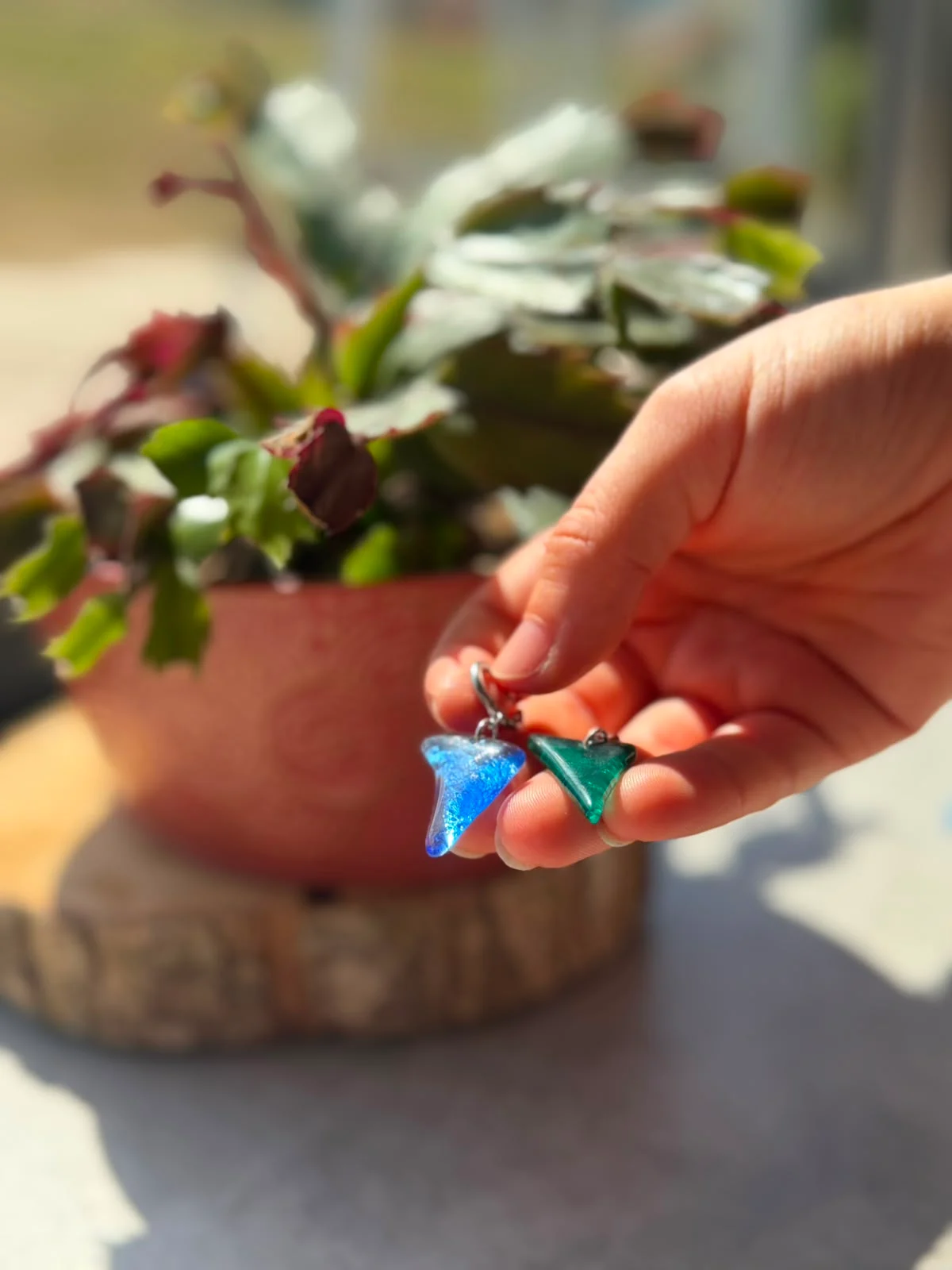 Main tenant deux boucles d’oreilles pendantes en verre bleu et vert devant une plante en pot.