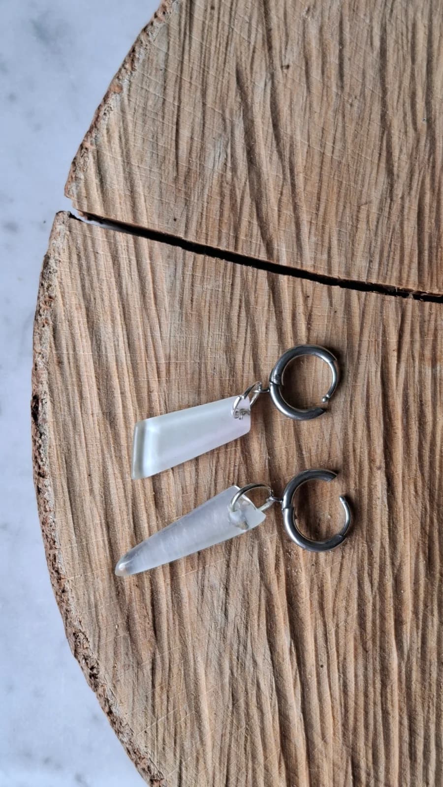Boucles d'oreilles asymétriques en verre blanc laiteux et anneaux en acier posées sur du bois.