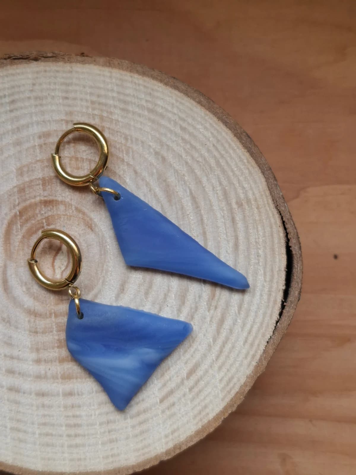 Boucles d'oreilles dormeuses dorées ornées de pièces géométriques asymétriques en verre bleu sur rondin de bois.