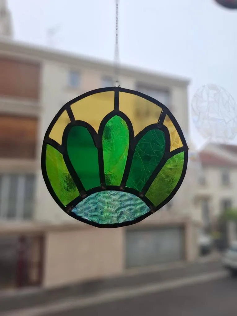 Vitrail rond suspendu aux motifs de pétales verts, verre jaune et bleu texturé.