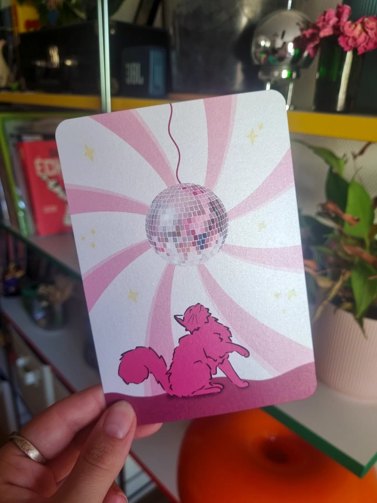 Carte illustrée avec un chat rose observant une boule à facettes disco sur un fond à rayons roses et blancs