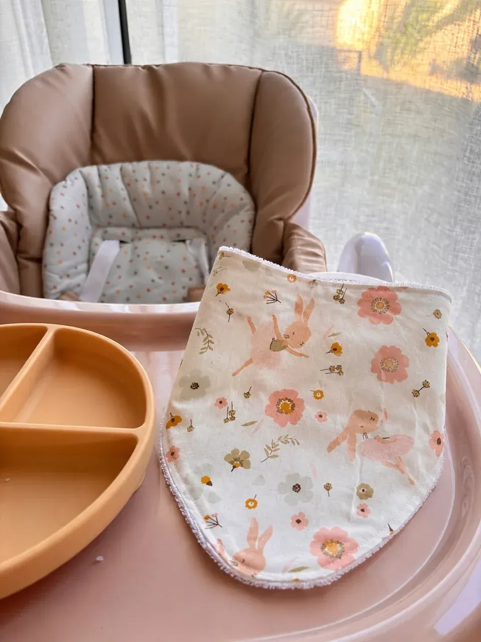 Bavoir bandana beige à motifs de petits lapins et fleurs roses posé sur le plateau d'une chaise haute.