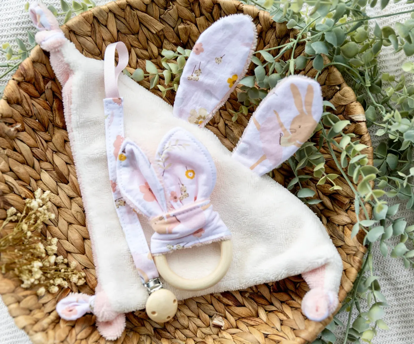 Doudou plat lapin écru et rose, anneau de dentition et attache-tétine fleuris dans un panier tressé