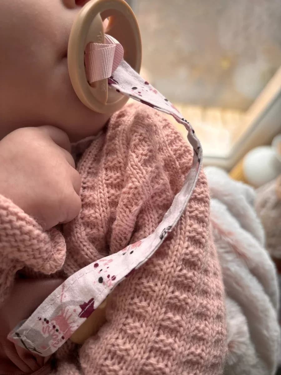 Attache-tétine en tissu à motifs avec clip rond en bois posée sur un plaid duveteux blanc.