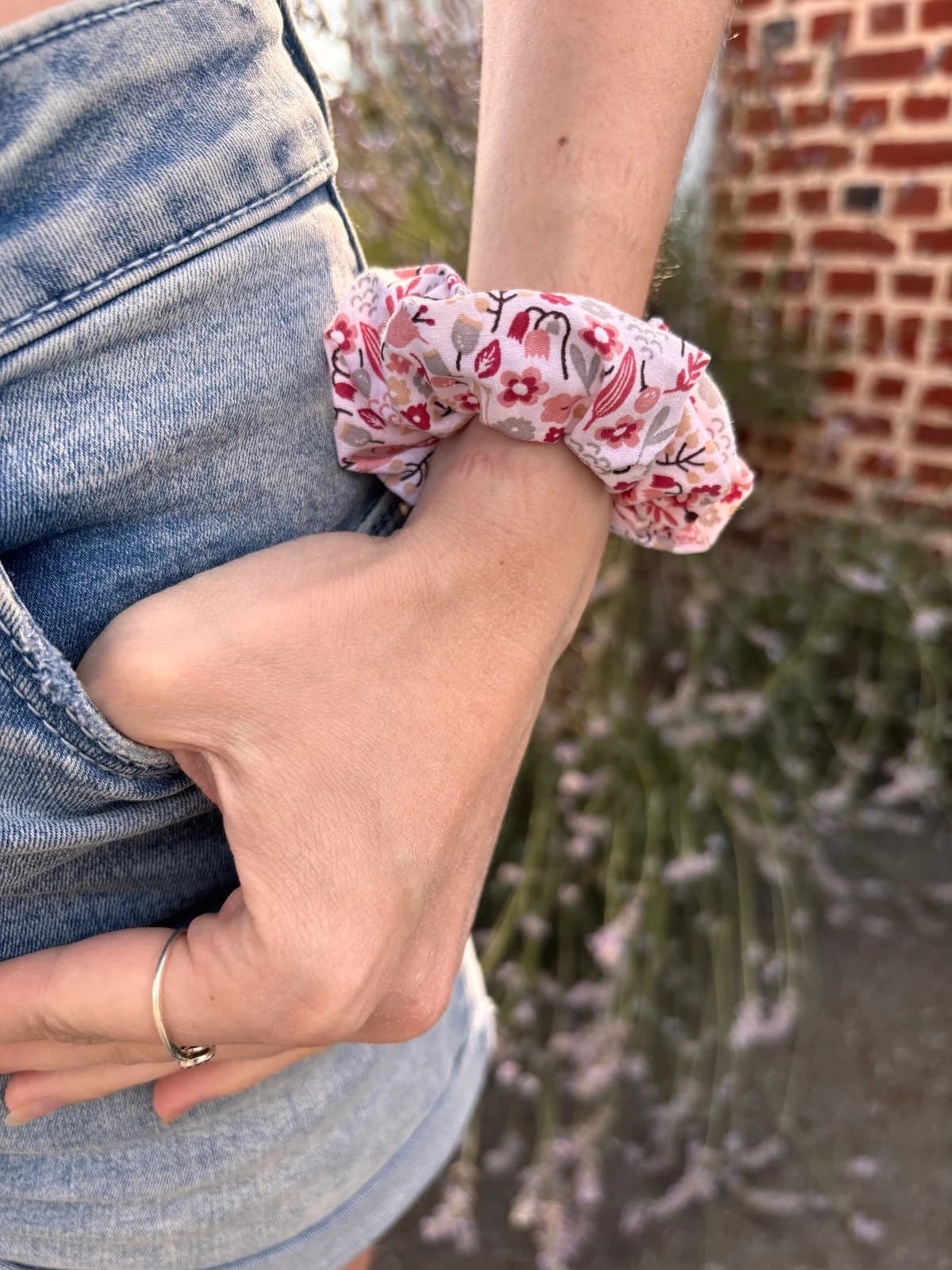 Chouchou en tissu à motifs floraux roses et bordeaux porté au poignet, main dans la poche d'un jean.