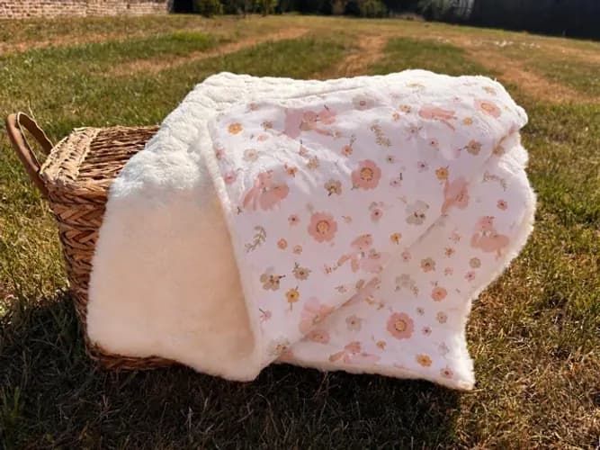 Couverture bébé pliée sur un panier en osier, avec un côté polaire écru et un côté en tissu à motifs floraux roses.