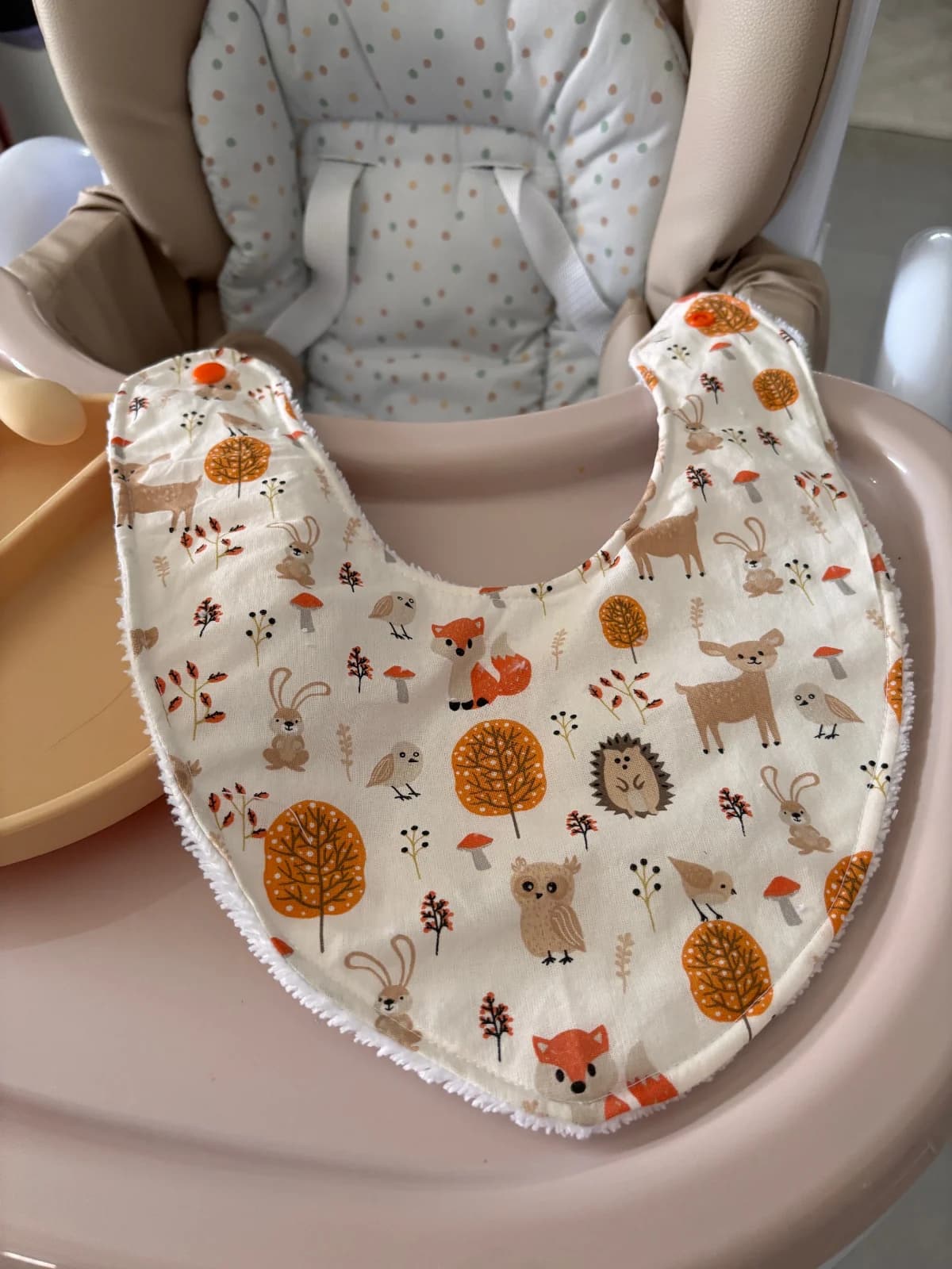 Bavoir bandana écru à motifs animaux d'automne, renard et arbres orangés, posé à plat sur une chaise haute.