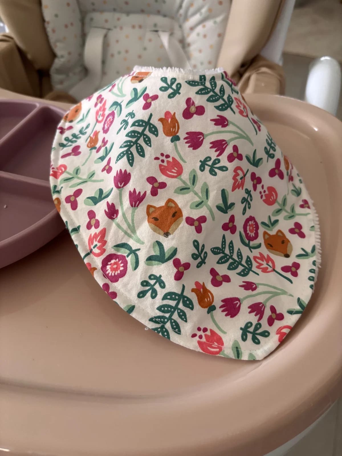 Bavoir bandana aux motifs floraux et têtes de renard, posé sur une tablette de chaise haute.