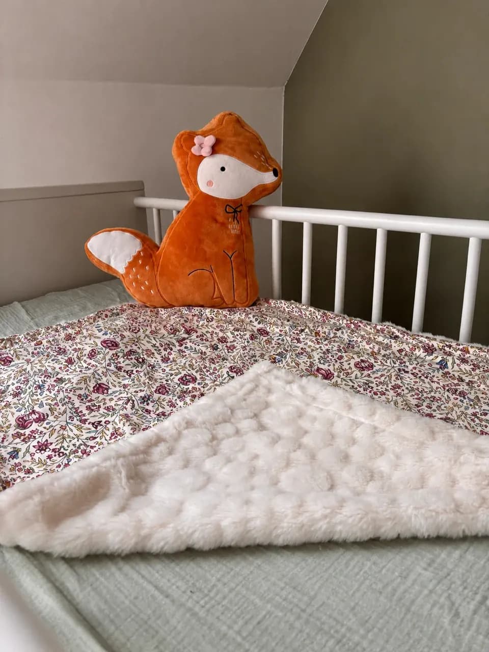 Couverture bébé à motif fleuri et polaire texturée blanche posée sur un lit d'enfant avec coussin renard