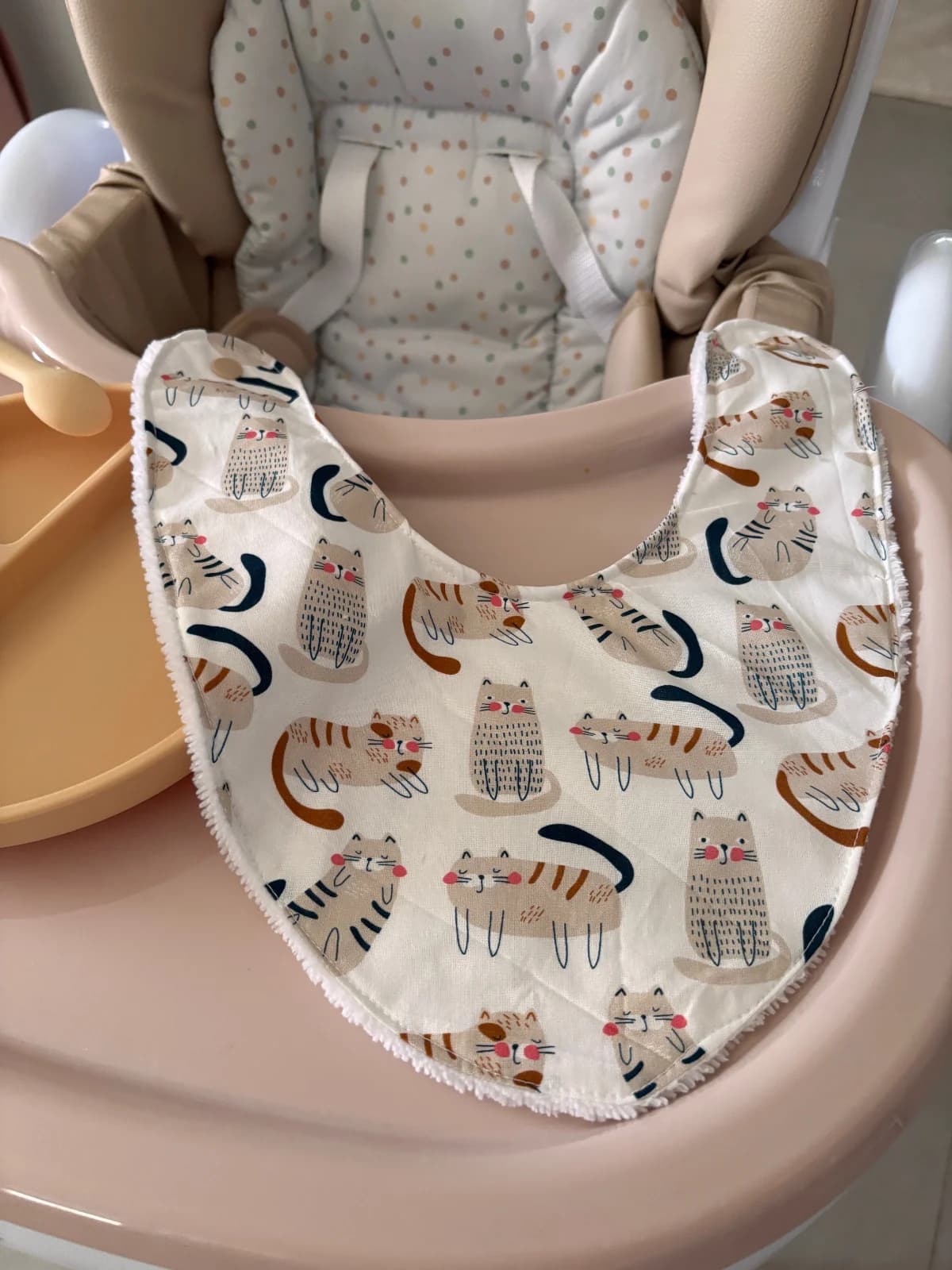 Bavoir bandana blanc imprimé de chats beiges posé à plat sur le plateau d'une chaise haute pour bébé.