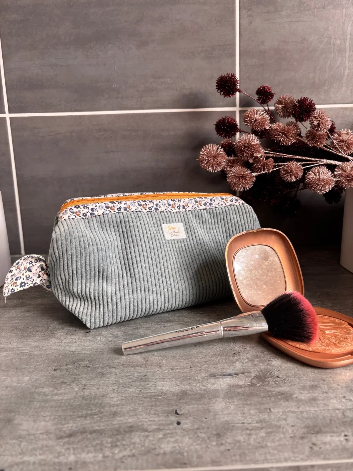 Trousse fermée en velours côtelé vert d'eau et tissu fleuri posée à côté d'un pinceau de maquillage.
