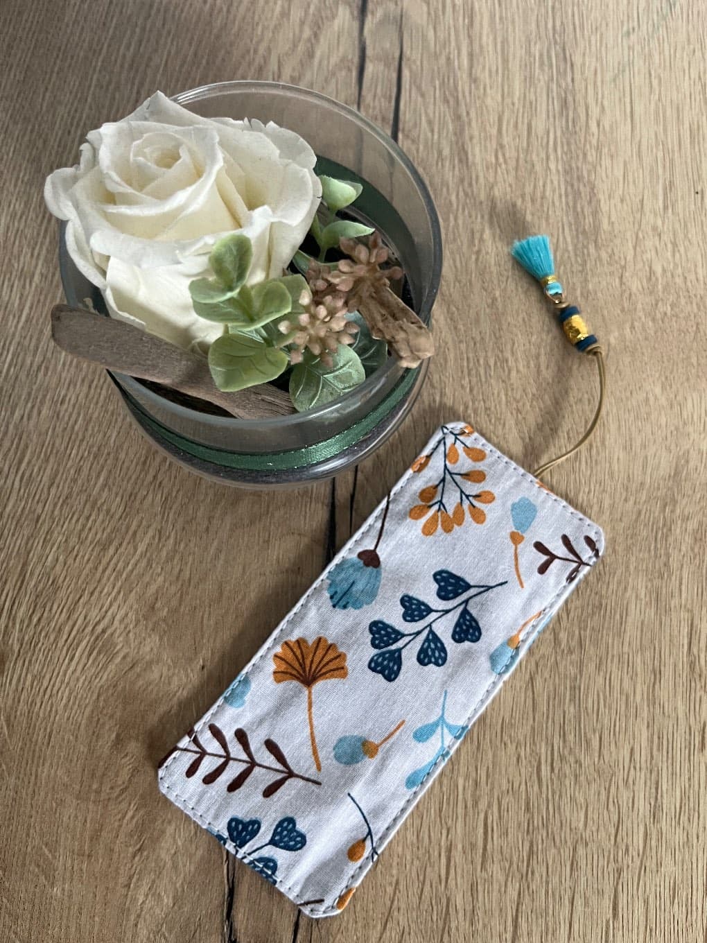 Marque-page rectangulaire en tissu à motifs végétaux bleu et moutarde avec pompon turquoise.