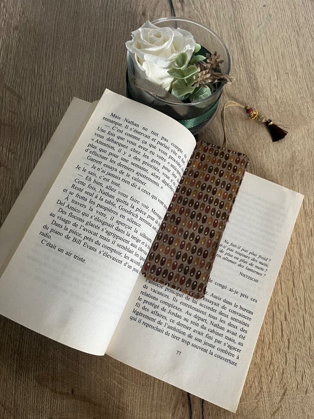Marque-page en tissu à motifs marron et ocre avec cordon et pompon marron, posé sur une table en bois.