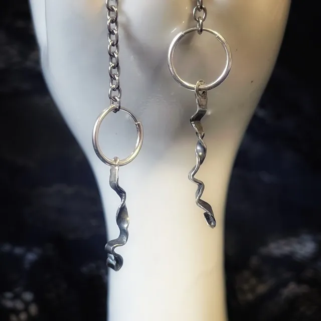 Paire de créoles en argent avec pendentifs ondulés façon ruban sur support blanc.