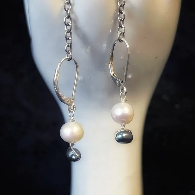 Boucles d'oreilles pendantes à perles blanches et grises suspendues à une main sculptée sur fond dentelle.