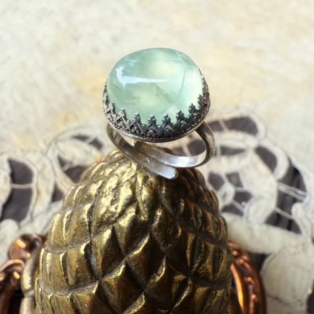 Bague en argent sertie d'un cabochon vert clair, posée sur un support métallique doré façon pomme de pin