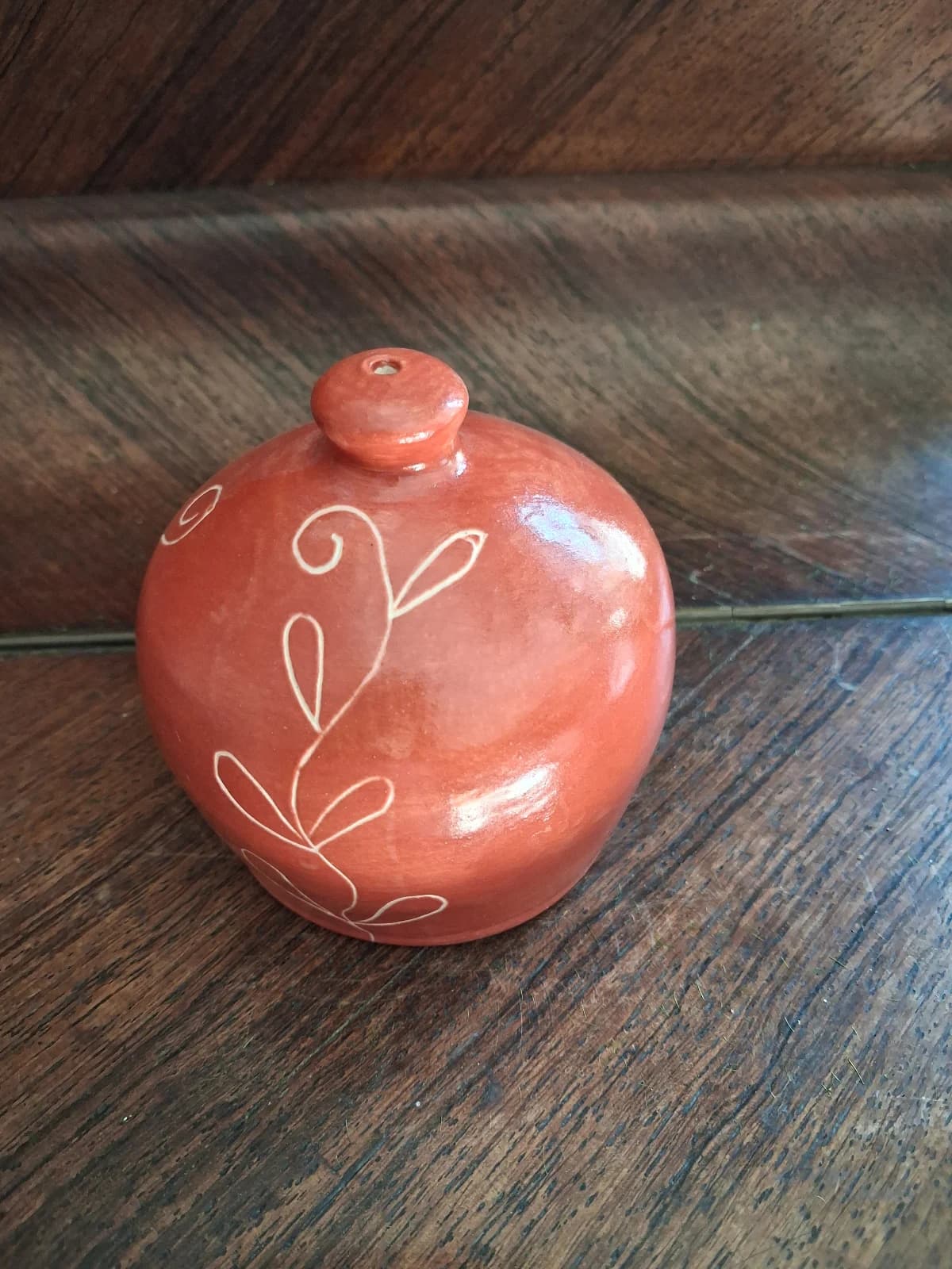 Arrosoir-main sphérique en céramique rouge brique orné d'un dessin végétal blanc, sur un meuble en bois.