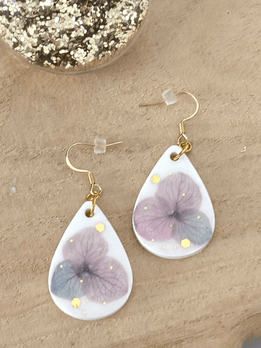 Boucles d'oreilles pendantes blanches en goutte avec fleur mauve séchée et touches dorées sur fond bois.