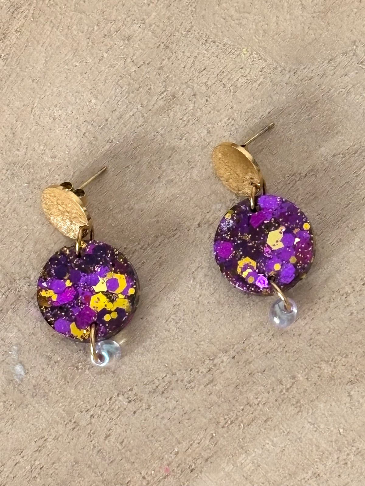 Boucles d'oreilles dorées avec médaillons ronds violets à paillettes posées sur fond bois.