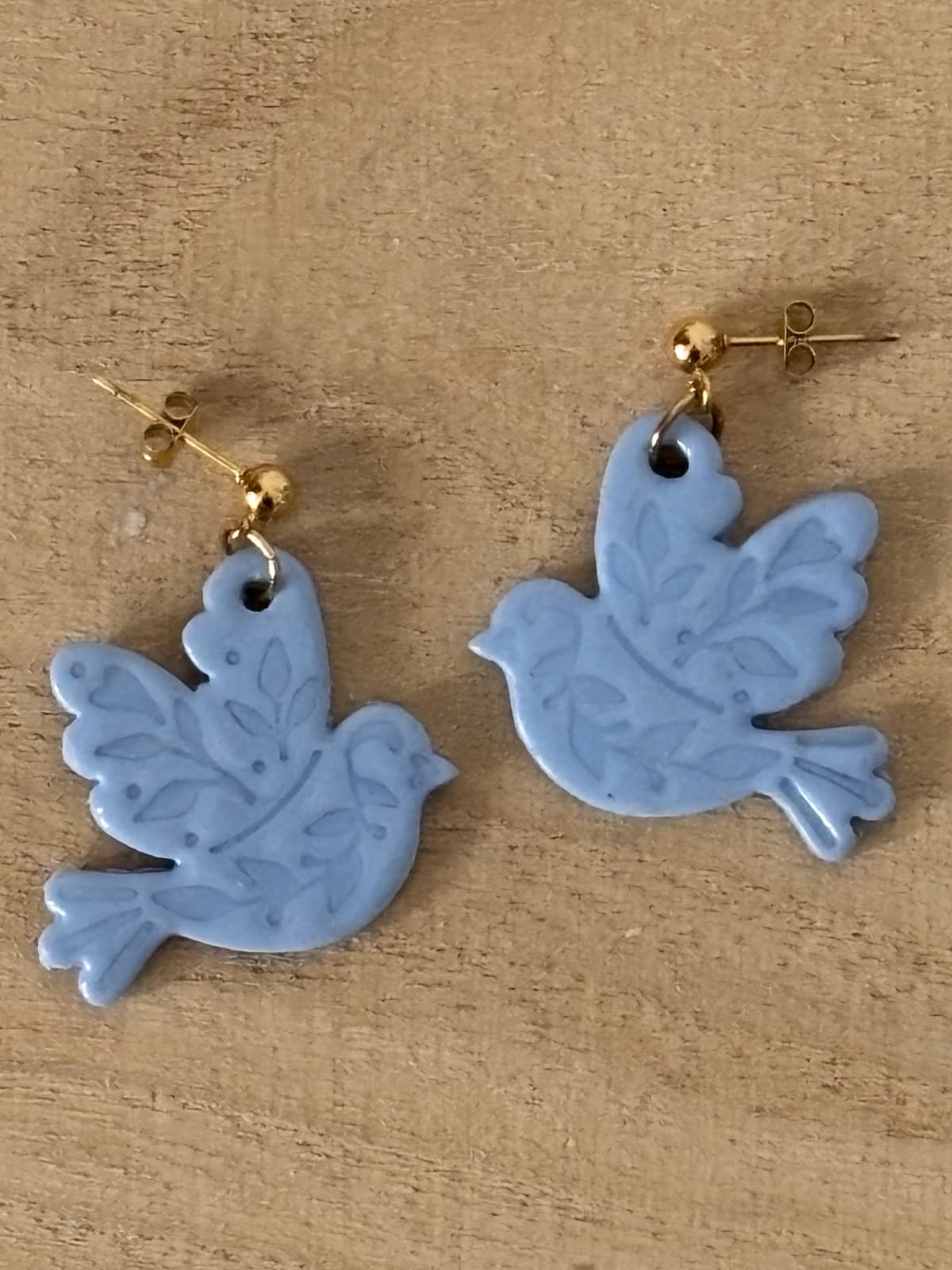 Boucles d’oreilles bleu gris en forme d’oiseaux gravés avec attaches dorées, posées sur du bois.