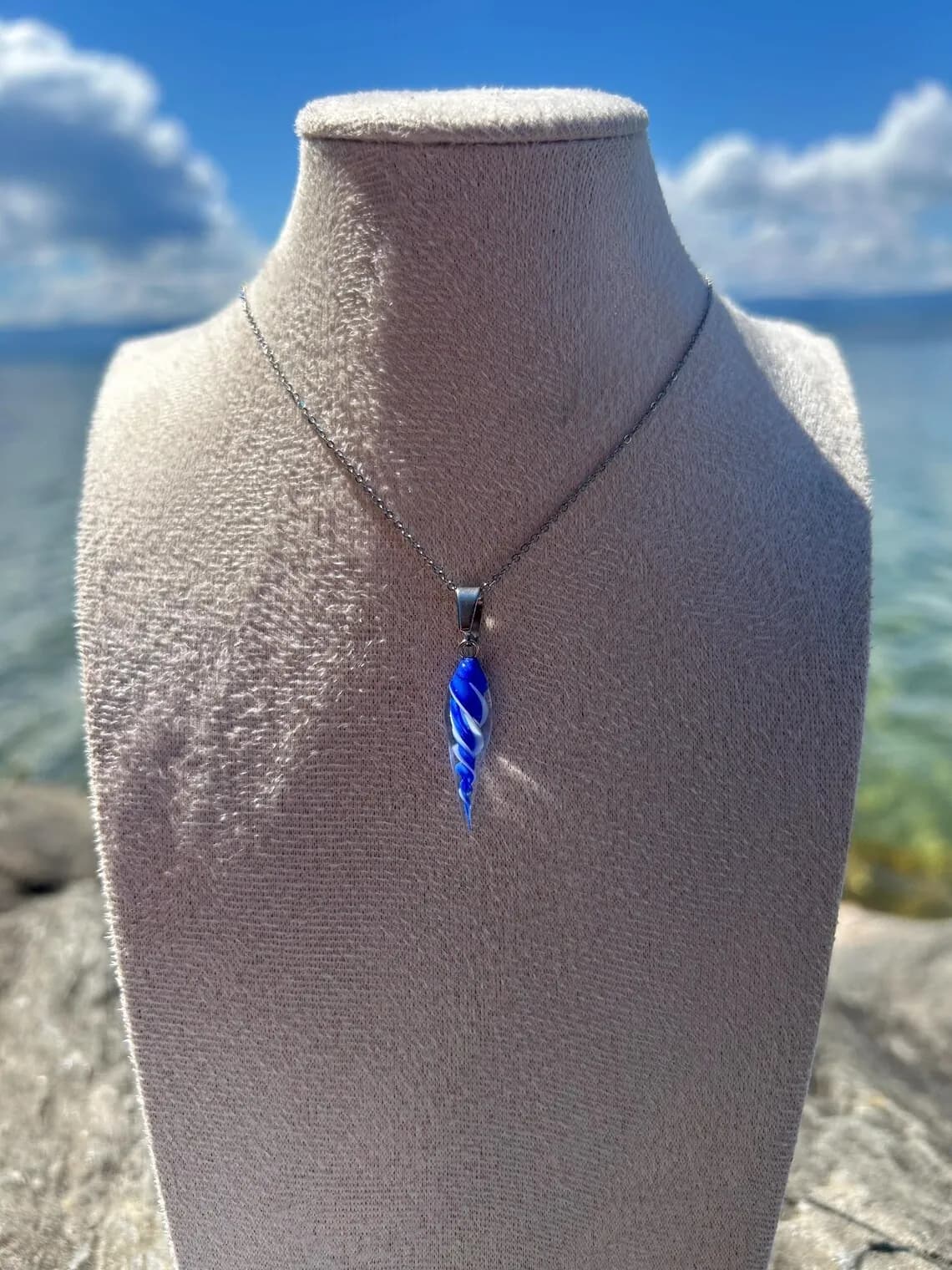 Pendentif coquillage torsadé en verre filé bleu et blanc sur fine chaîne, porté sur buste devant l'eau.