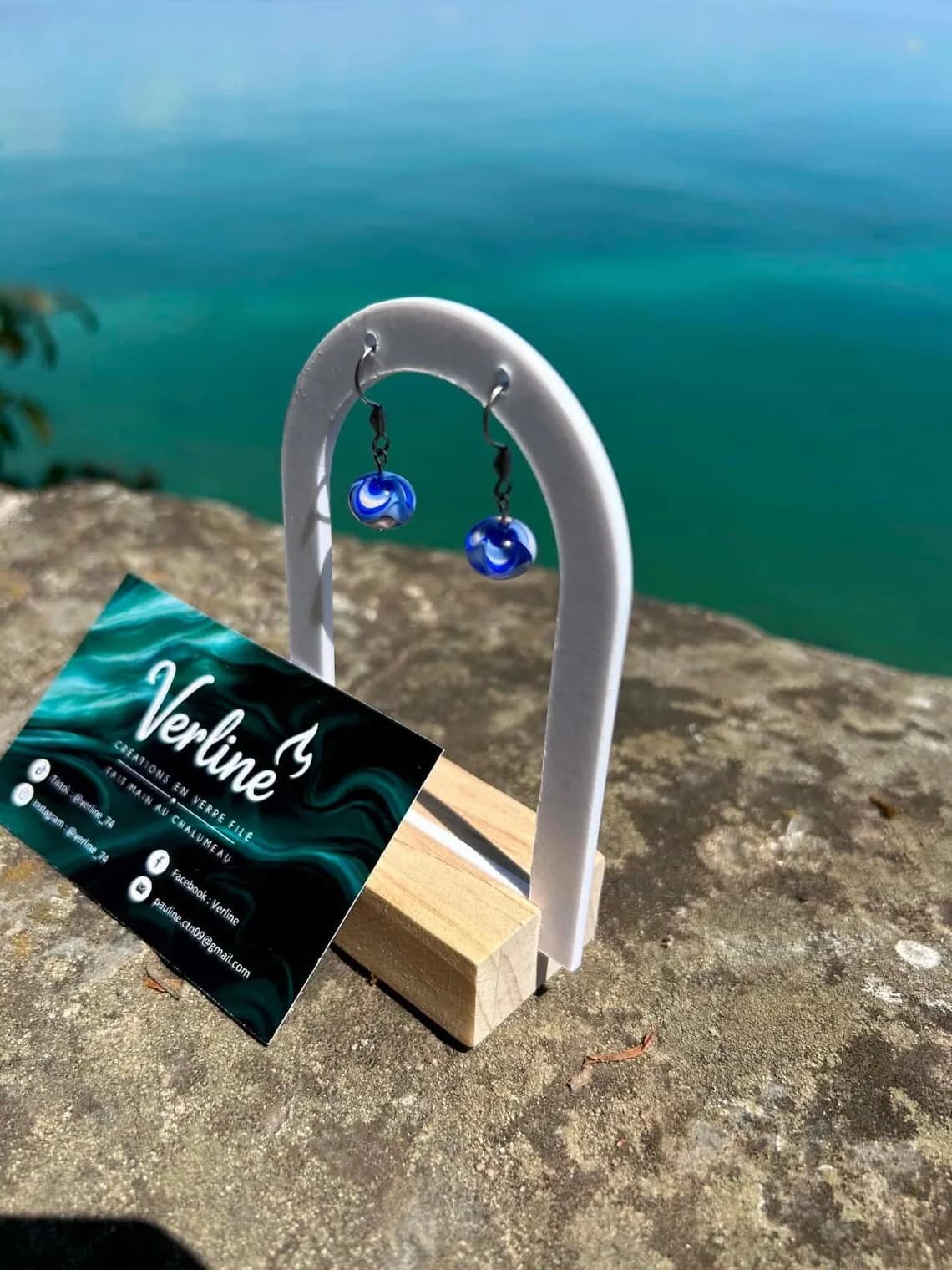 Paire de boucles d'oreilles en verre bleu et blanc sur un support blanc en arche, face à la mer.