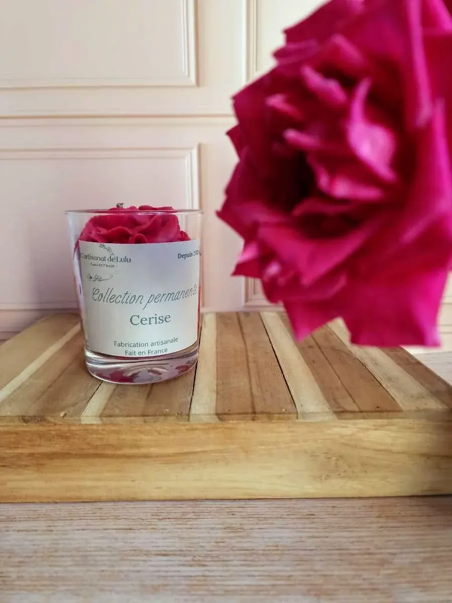 Bougie artisanale en pot de verre étiqueté Cerise, posée sur un plateau en bois devant une fleur rose.