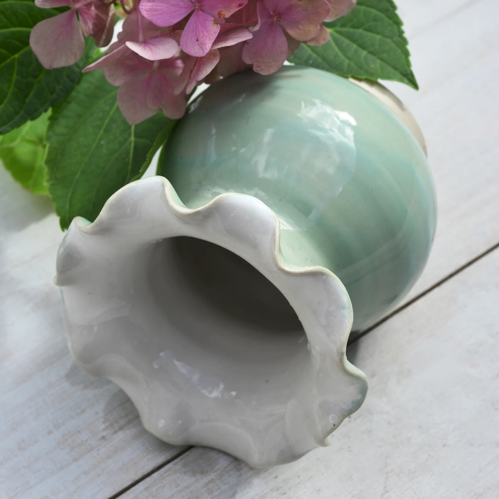 Vase boule en céramique vert d'eau à col évasé ondulé blanc, posé près de fleurs roses sur fond bois.