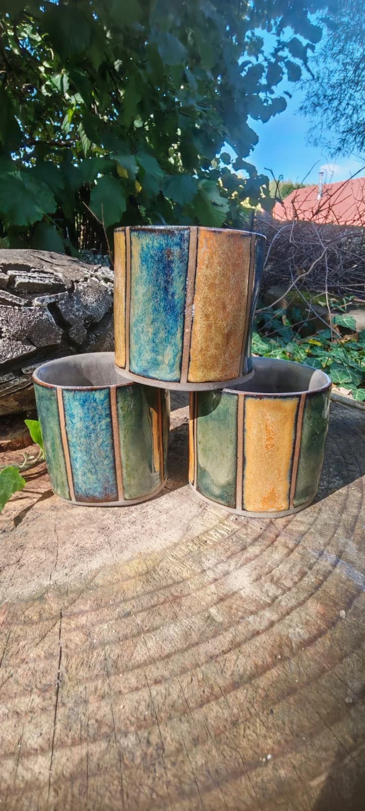 Trois gobelets en céramique aux bandes verticales bleu, vert et ocre posés sur une souche de bois.