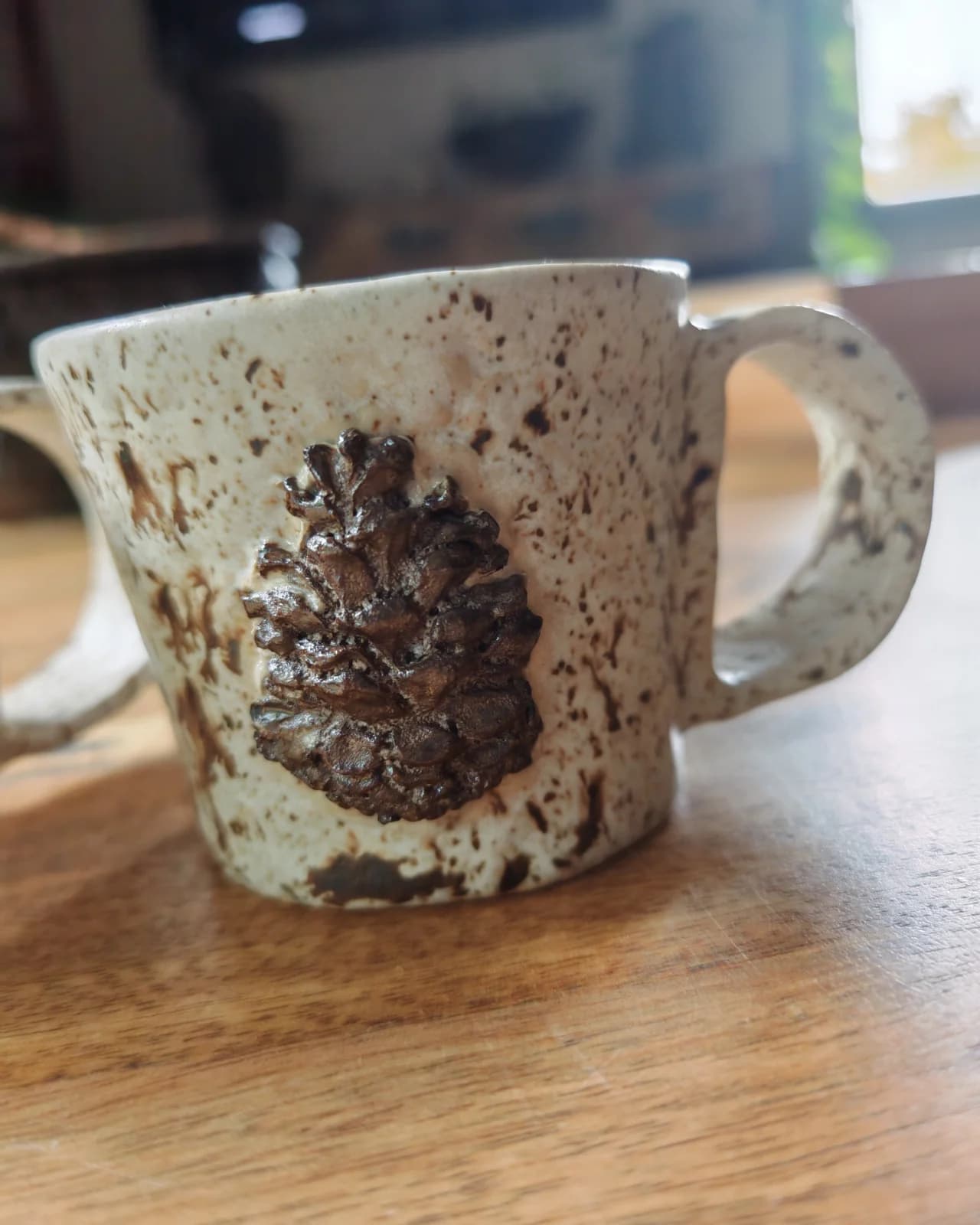 Gros plan sur une tasse en céramique claire mouchetée ornée d'une pomme de pin en relief.