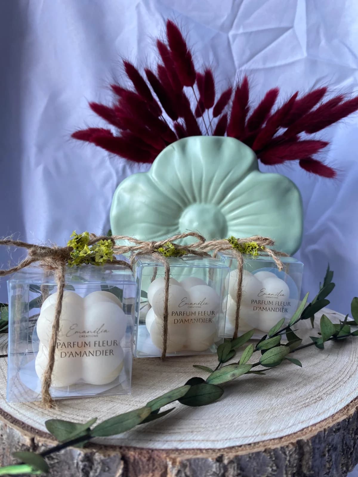 Trois fondants parfumés blancs en boîte transparente avec ficelle et fleurs séchées sur rondin de bois.