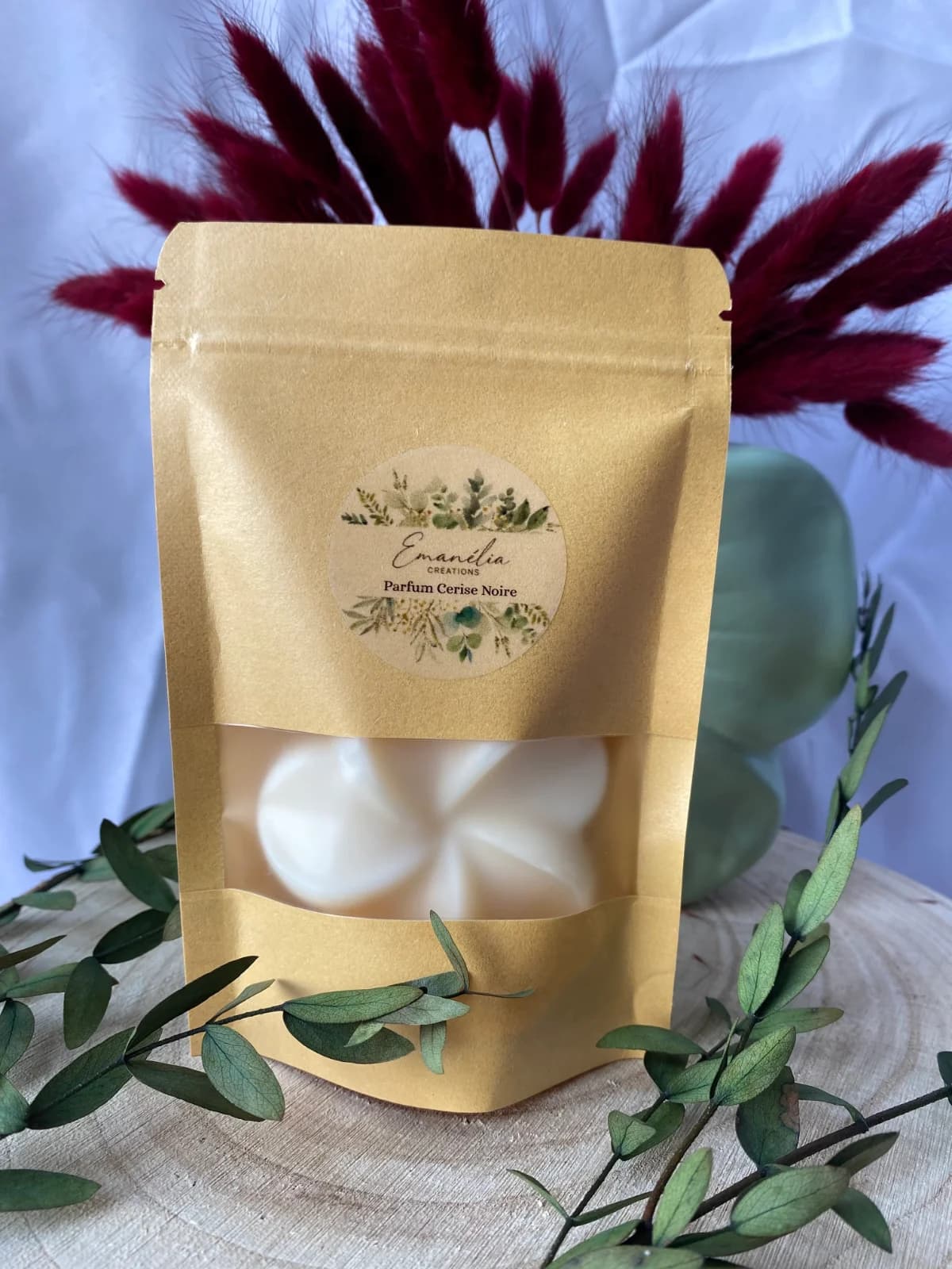 Fondant blanc en sachet kraft posé sur un rondin de bois avec feuillage et fleurs séchées bordeaux.